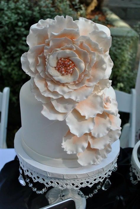 White cake with large, detailed white fondant flowers on top, adorned with small pearl-like decorations. The cake sits on a lace-trimmed stand with crystal accents.