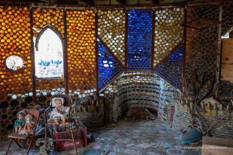 Interior view of a room with walls made from colorful glass bottles, featuring old dolls and toys displayed on the left side and a stone floor.