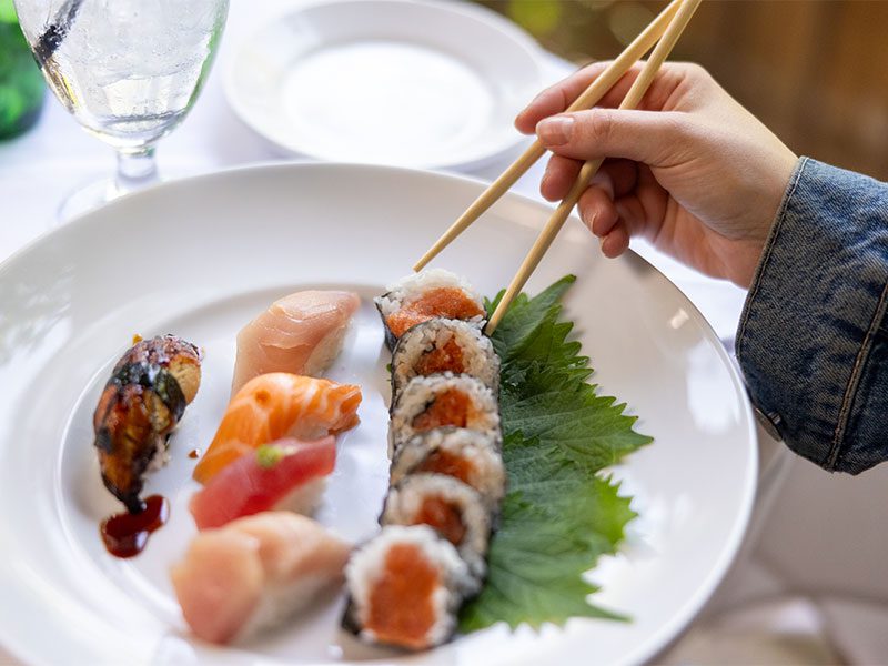 A hand using chopsticks picks up a sushi roll from a plate with various sushi pieces, garnished with a green leaf. A glass of water is in the background.